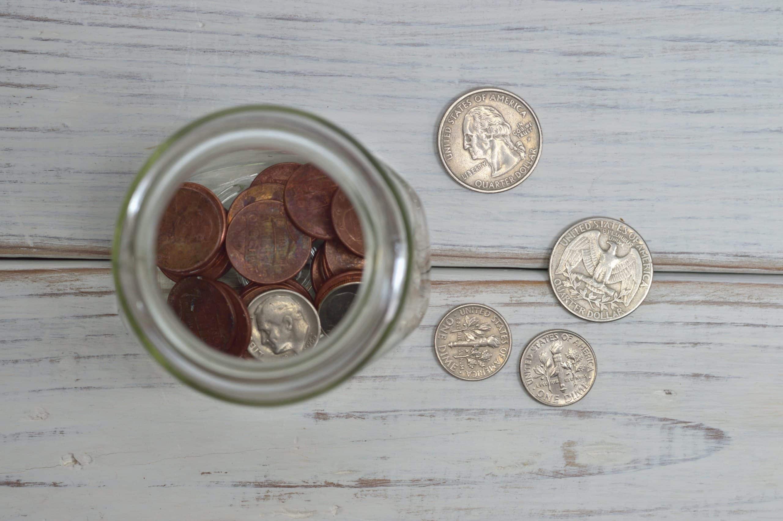 A jar filled with pennies viewed from above, placed on a wooden surface. Nearby, there are several quarters and dimes scattered on the table.
