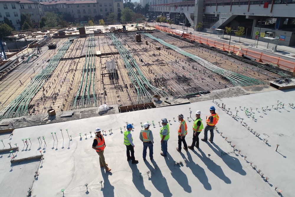 A group of construction workers in safety vests and helmets stand on a concrete slab at a construction site, surrounded by equipment and wiring, with a building and trees in the background.