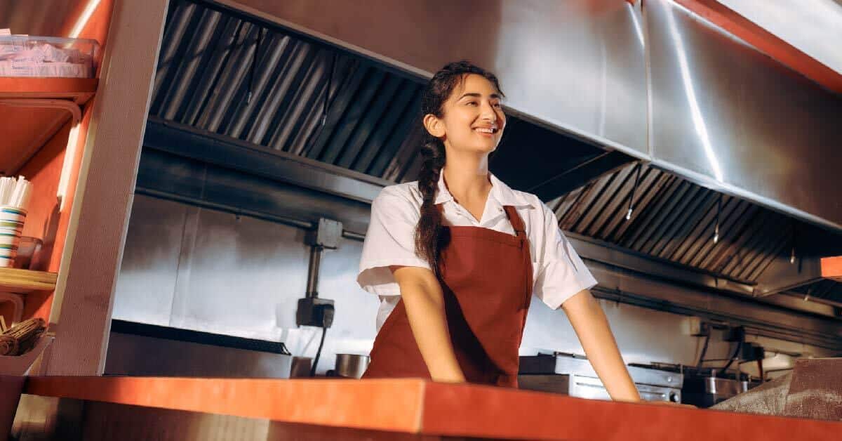 A person wearing a white shirt and red apron smiles while standing in a commercial kitchen.