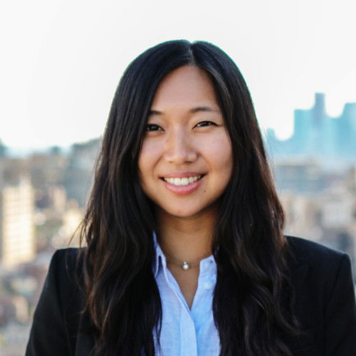 A person with long dark hair is standing outdoors, wearing a black blazer and a light blue shirt, smiling with a blurred cityscape in the background.
