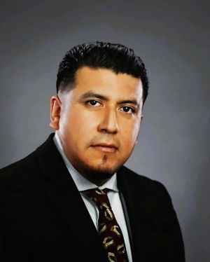 A man with short dark hair, trimmed beard, and mustache is wearing a dark suit, white shirt, and patterned tie, posing against a plain gray background.