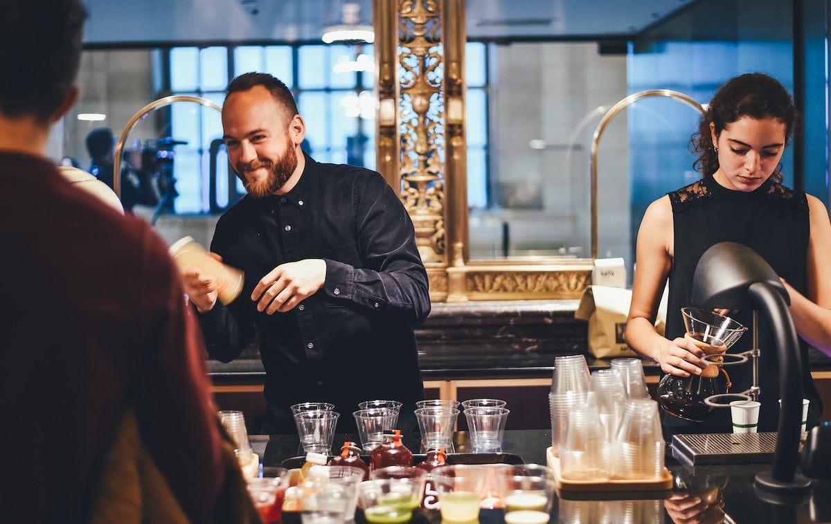 Two baristas, a man and a woman, are preparing drinks behind a counter in a café. The man is shaking a cocktail shaker while the woman is focused on a coffee machine.