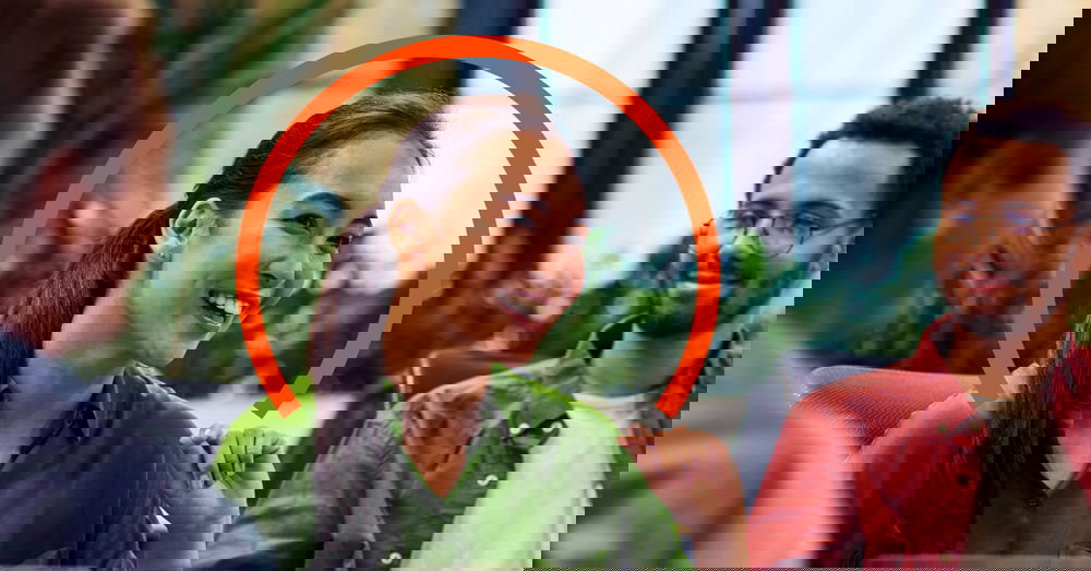 A woman in a green shirt smiles during a group meeting, with an orange semicircle graphic behind her head. Two other people are seated nearby.