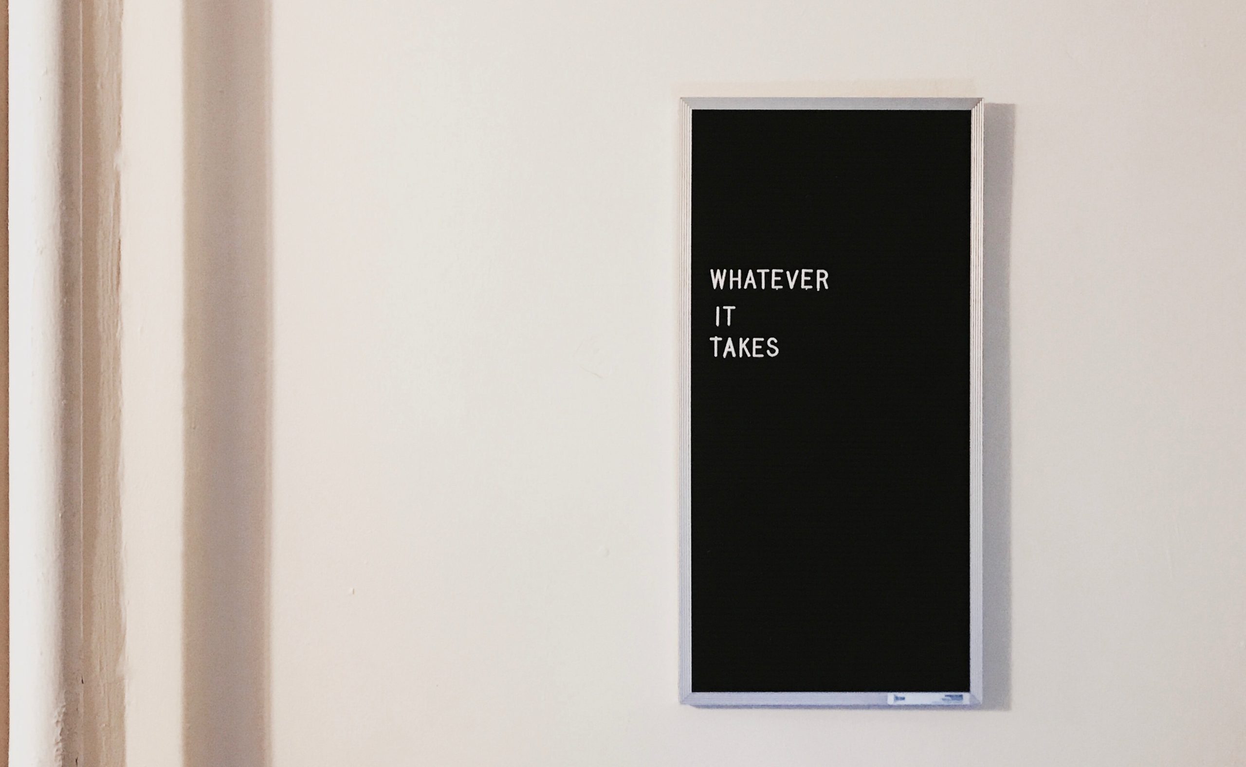 A black sign with white text that reads "WHATEVER IT TAKES" hangs on a plain white wall.