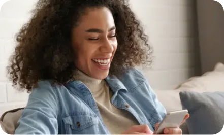 A person with curly hair and wearing a denim jacket smiles while looking at a smartphone.