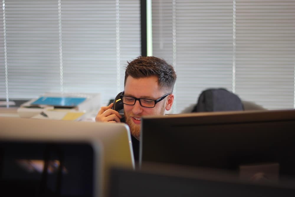 A man with glasses and a beard is talking on the phone in an office with blinds. Multiple computer monitors and office equipment are visible.