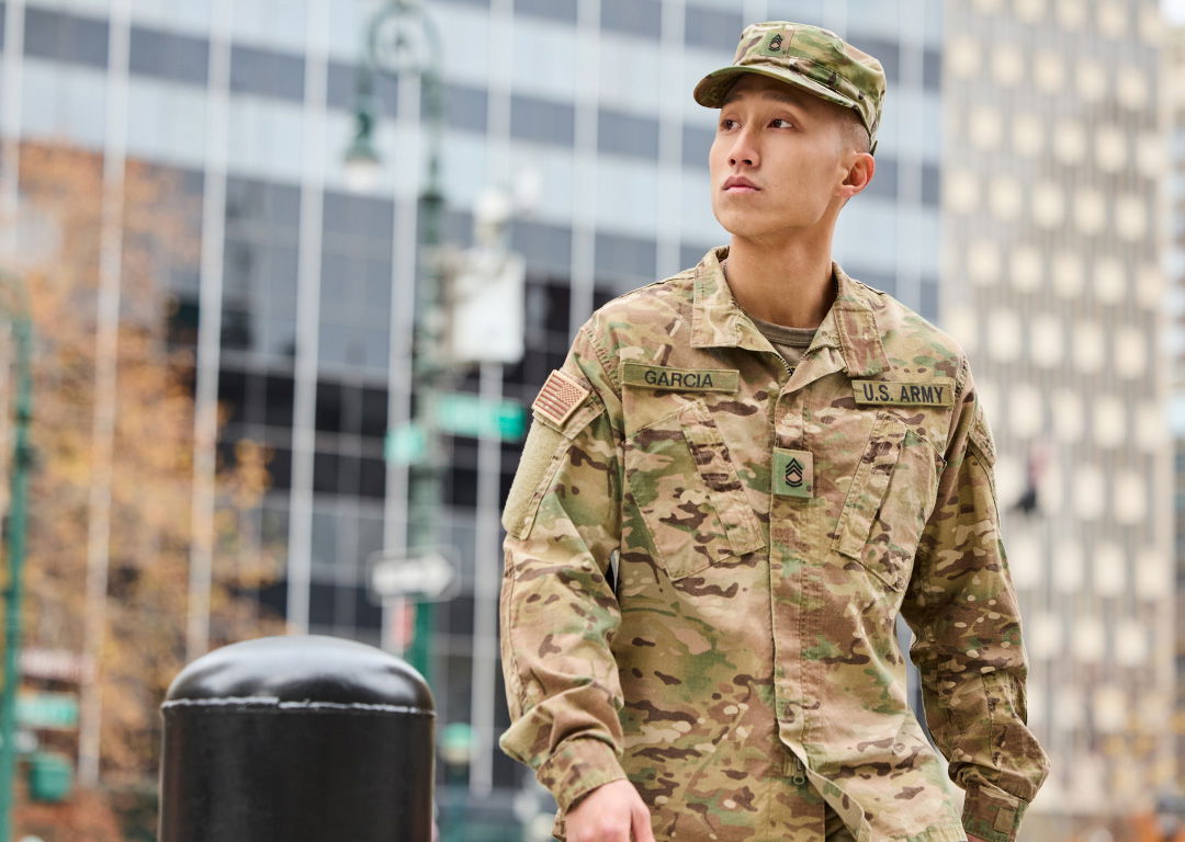 A person in U.S. Army camouflage uniform and cap stands outdoors in an urban setting with glass buildings in the background.