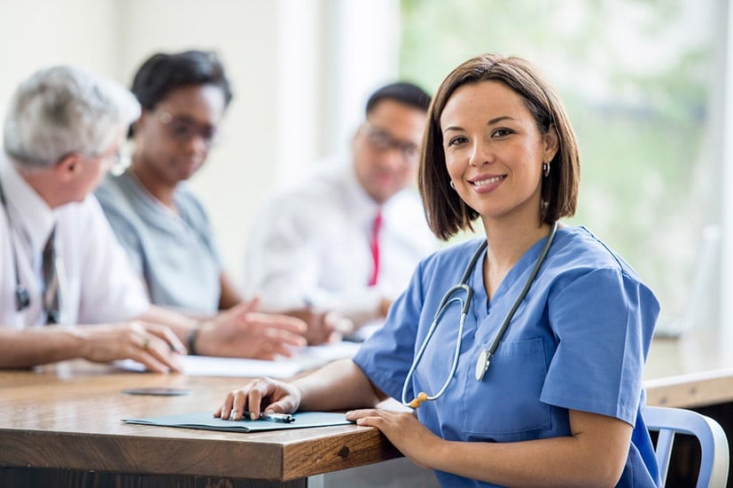 A healthcare professional in blue scrubs and a stethoscope smiles at the camera, with colleagues in discussion in the background.