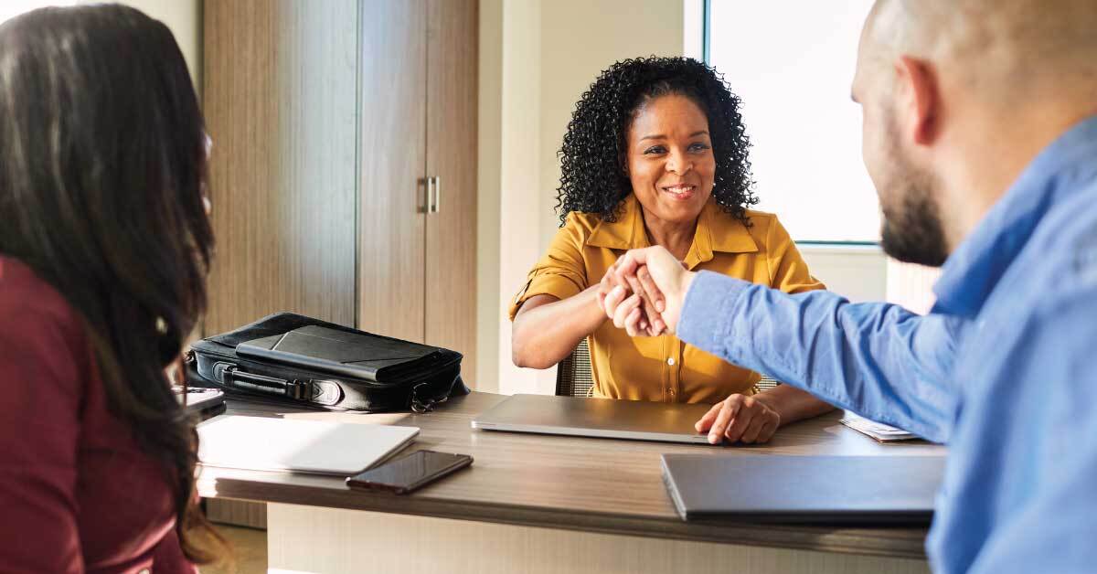 A woman in a yellow shirt shakes hands with a man in a blue shirt while another woman sits nearby in an office setting. A black bag is on the table.
