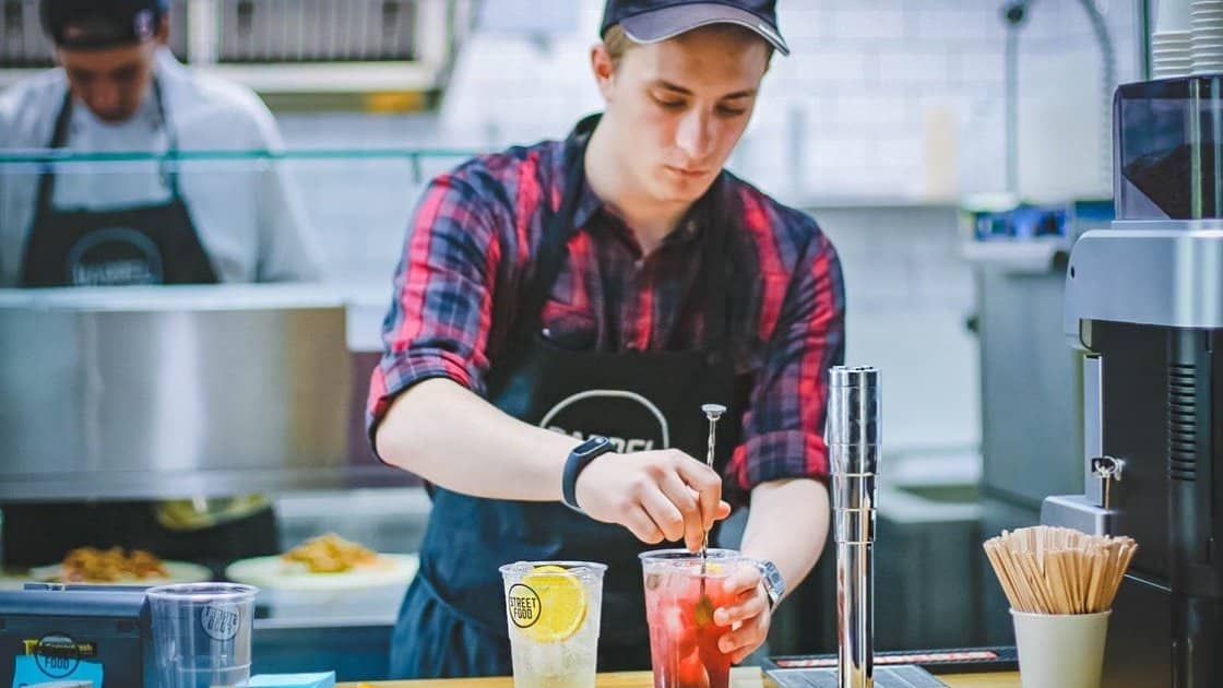 Man in a plaid shirt and cap prepares a red beverage at a counter, with another man in the background cooking. Various drink-making utensils and ingredients are visible on the counter.