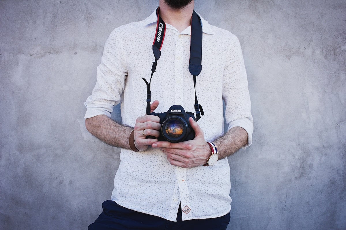 A person wearing a white shirt holds a Canon camera with both hands, against a gray concrete wall background.