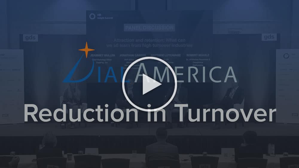 A conference slide with the title "Reduction in Turnover" and a play button in the center. The background shows a panel discussion with participants sitting on stage.