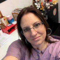 Woman with glasses and shoulder-length brown hair wearing a purple top, sitting at a desk with office supplies and files in the background.