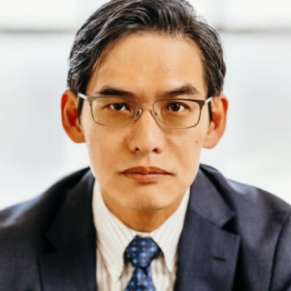 A man wearing glasses and a suit with a blue tie looks directly at the camera with a neutral expression.