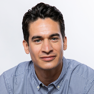 A man with dark hair wearing a blue and white checkered button-down shirt looks directly at the camera against a plain background.