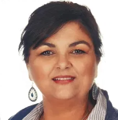 A woman with dark hair, wearing blue and white earrings, a striped collared shirt, and a blue jacket, smiles at the camera against a plain white background.