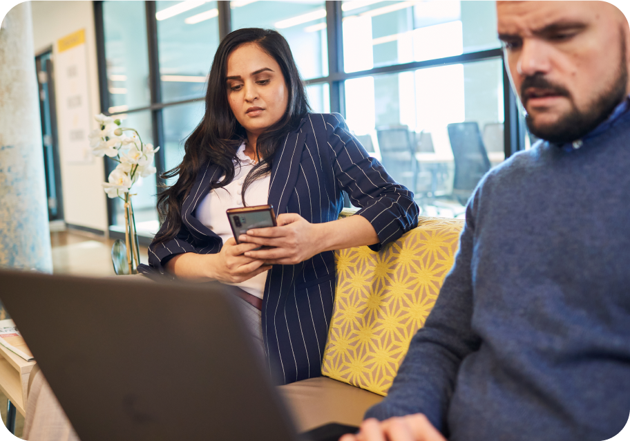 Two people sit on a yellow couch; one woman looks at her phone while a man works on a laptop. Office setting with large windows in the background.