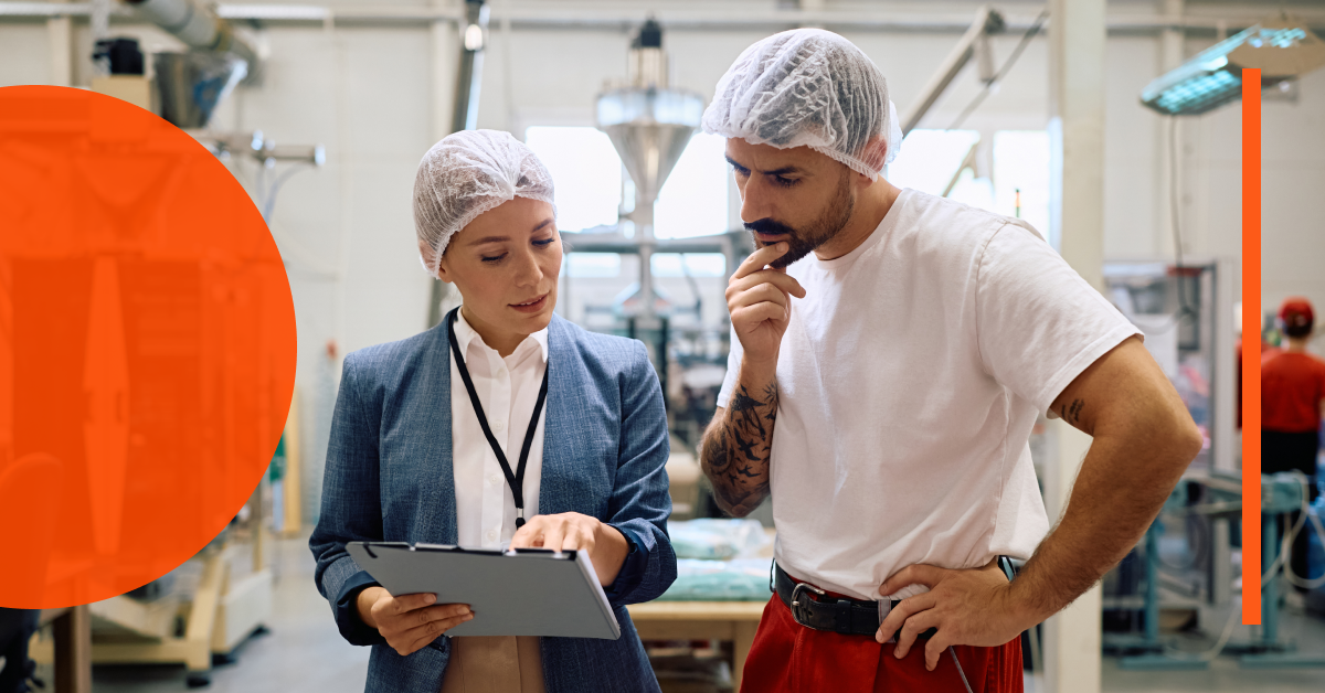 Two workers in hairnets, one in business attire and one in casual clothes, review information on a clipboard inside a factory or industrial setting.
