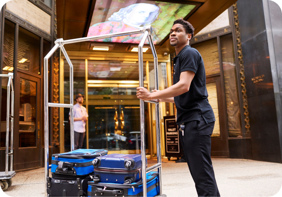 A hotel staff member stands outside an entrance, pushing a luggage cart loaded with blue suitcases. A valet parking sign and a digital display are visible in the background.
