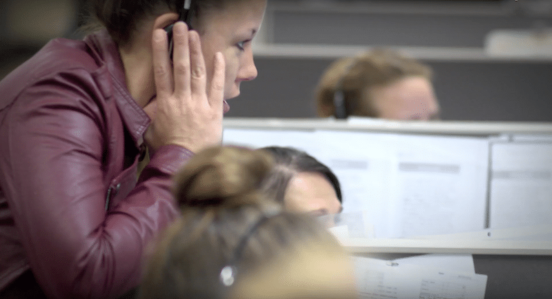 A woman wearing a headset and a maroon jacket is working at a desk in a call center, with other people working in the background.