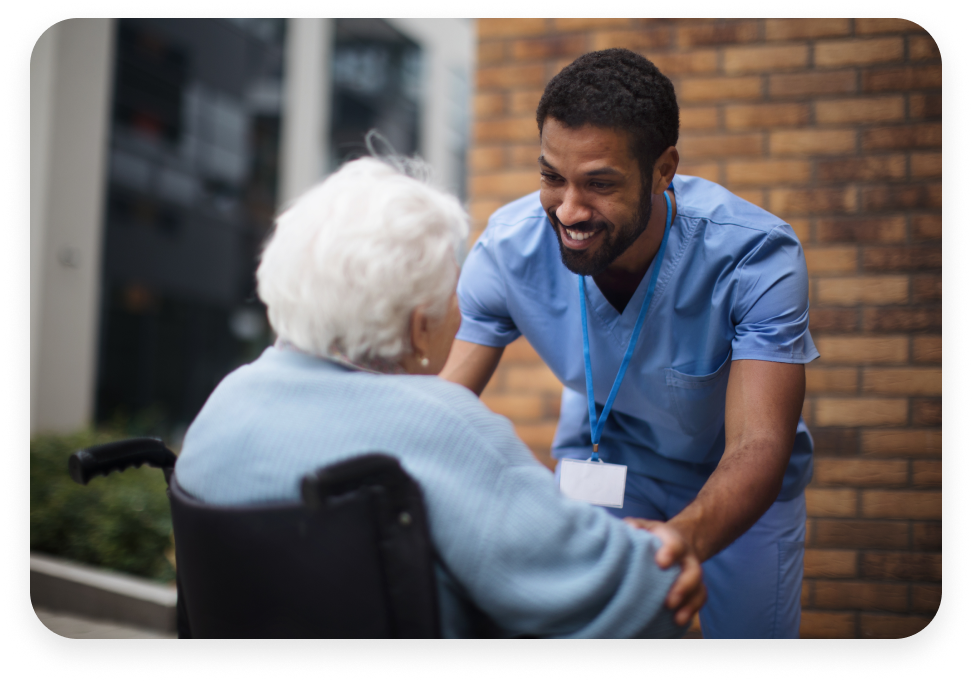 A healthcare worker in blue scrubs and a name badge smiles at an elderly person in a wheelchair outside a building, holding their hands.