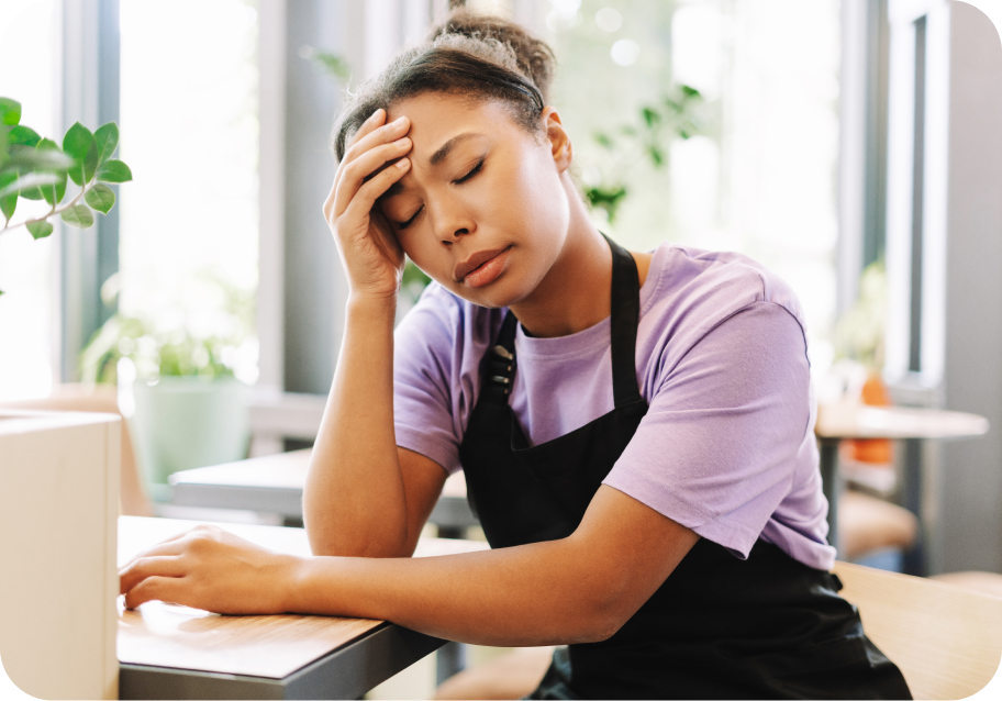 A person wearing a purple shirt and black apron sits at a table, resting their head in their hand, appearing tired.