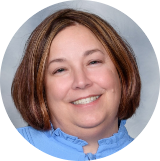 A woman with shoulder-length brown hair smiles at the camera. She is wearing a light blue shirt and has a light-colored background.