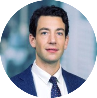 A man with short dark hair, wearing a blue suit, white shirt, and polka dot tie, poses in front of a blurred office background.