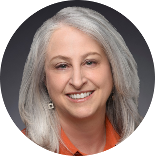 Portrait of a smiling woman with long, silver hair, wearing an orange top and square earrings, against a dark background.