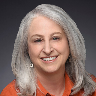 A woman with shoulder-length gray hair, wearing an orange collared shirt and geometric earrings, smiles at the camera against a dark background.