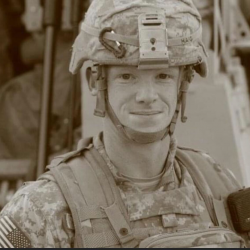 A soldier in full combat gear, including a helmet and body armor, stands and faces the camera with a neutral expression. An American flag patch is visible on their uniform.