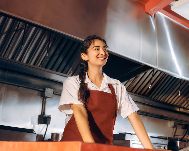 A person wearing a white shirt and brown apron stands behind a counter in a commercial kitchen, smiling.