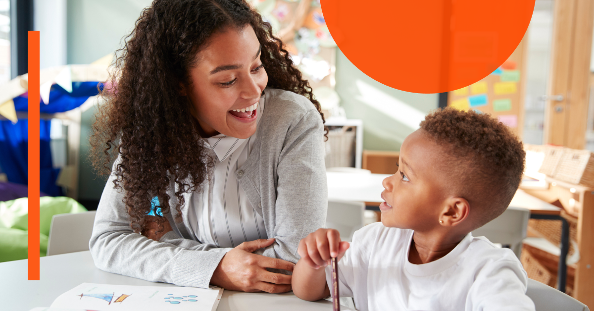 An adult and a child sit at a table in a classroom, smiling and talking to each other. Papers and drawings are spread out on the table.