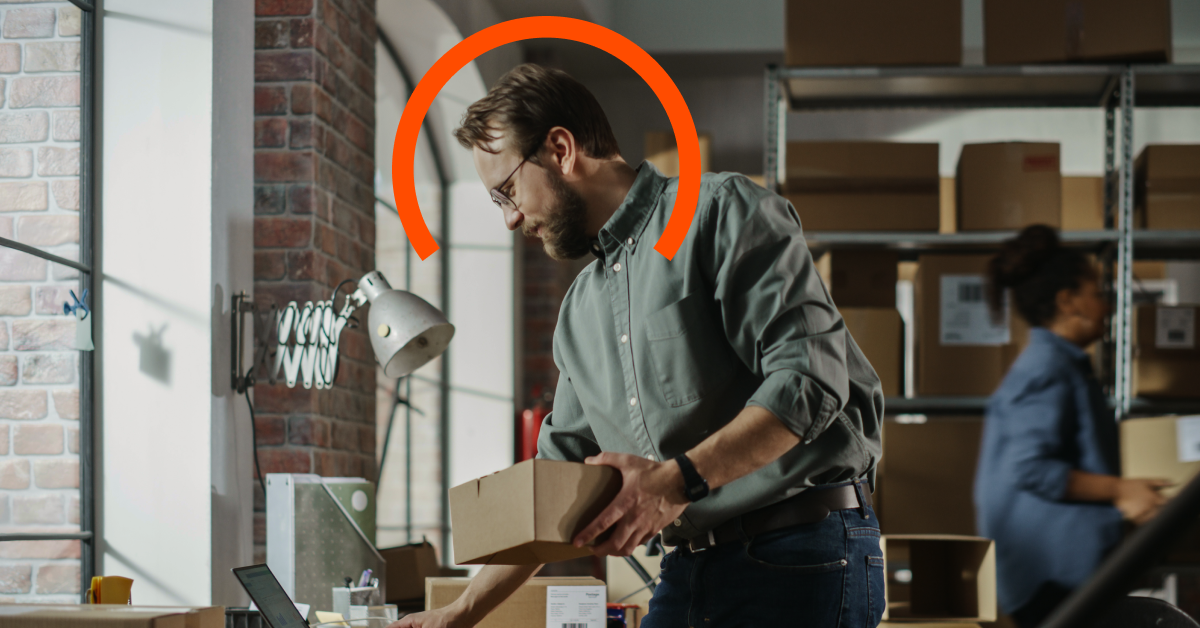 A man wearing glasses stands at a desk, holding a cardboard box and looking at a laptop in a warehouse or shipping office. An orange semi-circle is drawn above his head.