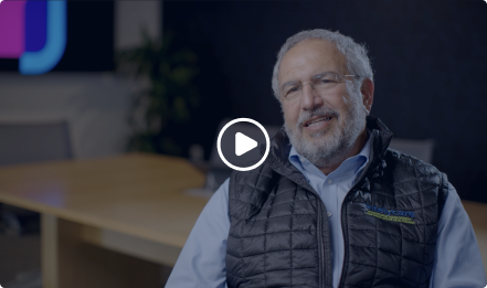 A man with gray hair and glasses, wearing a black quilted vest over a blue shirt, sits and smiles in an office setting. A play button overlay indicates this is a video thumbnail.