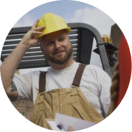 A construction worker in a yellow hard hat and beige overalls smiles while standing outside, holding a piece of paper.