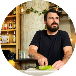 A man with a beard and tattoos slices fruits in a modern kitchen with shelves of bottles behind him.